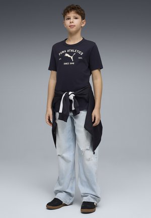 Granatowy bawełniany T-shirt z białym napisem i logo, w połączeniu z jasnoniebieskimi, podniszczonymi dżinsami. Czarne trampki z brązowymi podeszwami. Bluza zawiązana w talii.