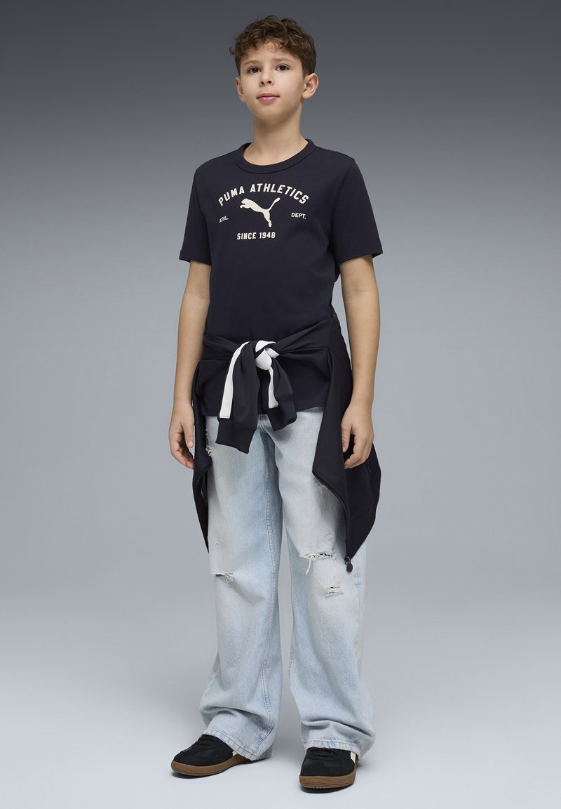 Tamsiai mėlynas medvilninis marškinėlis su baltais užrašais ir logotipu, derinamas su šviesiai mėlynomis, pažeistomis džinsais. Juodi sportbačiai su rudos spalvos padais. Džemperis surištas ties liemeniu.