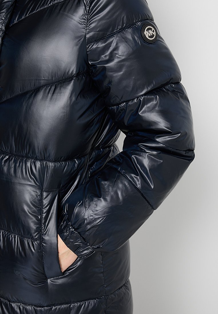 Veste matelassée bleu marine foncé avec une texture brillante et coussinée. Présente un logo rond sur la manche et des poches latérales. Coupe ajustée avec des poignets élastiques.