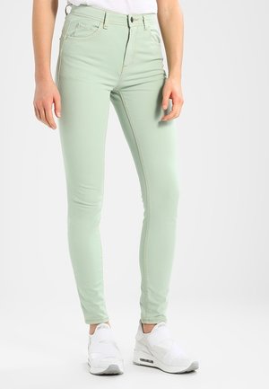 Slim-fit mintgroene broek gedragen met witte sneakers en een wit topje, weergegeven van taille tot voeten tegen een effen achtergrond.