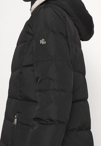 Doudoune noire à motif matelassé, avec une capuche doublée en peau lainée blanche, une fermeture éclair argentée et un logo brodé sur la manche.