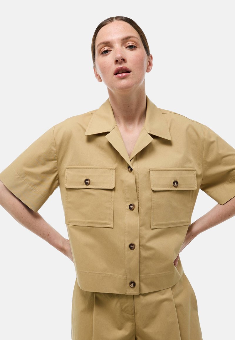 Chemise kaki à manches courtes avec un design court, deux poches avant et boutons marron. Texture lisse, col structuré, coupe ajustée.