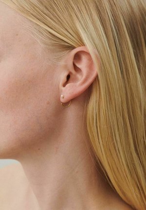 Lähedalt vaadates naise vasak kõrv, kus on väike kuldne rõngast kõrvarõngas ja väike pärlikujuline nael; blondid juuksed on kõrvast taha kinnitatud.