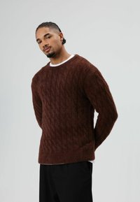 Bruine kabelgebreide trui met een gestructureerde afwerking, voorzien van een ronde halslijn en geribbelde manchetten, gedragen over een wit kledingstuk.