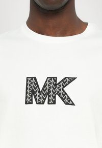 Hvit bomull t-skjorte med et stort, svart og grått brodert "MK"-logo med et geometrisk mønster. Enkel rund hals.