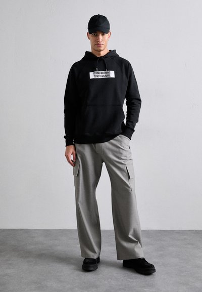 Un sweat à capuche noir avec un texte graphique blanc, associé à un pantalon cargo gris clair avec des poches. Le modèle porte une casquette noire et des chaussures noires épaisses.