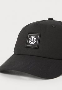 Gorra de béisbol negra hecha de un tejido texturizado que presenta un parche de logo cuadrado en blanco con elementos de diseño circulares y de árbol.