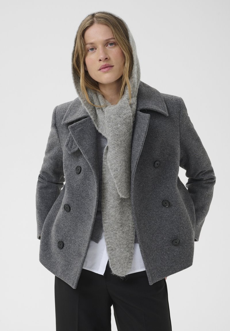 Femme portant un manteau gris croisé et une écharpe en tricot assortie sur une chemise blanche et un pantalon noir sur un fond uni.