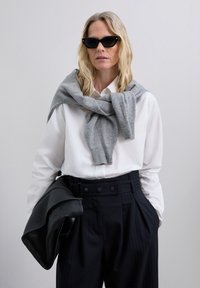 Grauer Wollschal über einem weißen Hemd mit Kragen, kombiniert mit hoch taillierten, gestreiften Hosen und einer schwarzen, strukturierten Ledertasche.