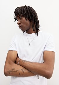 Bílé tričko, krátké rukávy, volný střih; osoba s copánky, která nosí stříbrný náhrdelník a náramek, vystavující tetování na pažích.