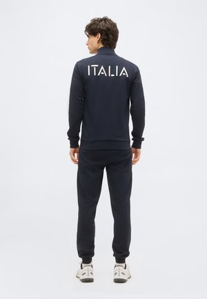 Hombre visto desde atrás, vestido con un chándal azul marino con la palabra "ITALIA" en letras blancas, rojas y verdes en la espalda, y zapatillas blancas.
