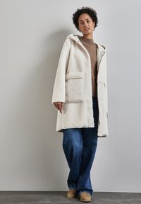 Crème-kleurige, gestructureerde fleecejas met capuchon, voorkeurszakken, open ontwerp, gedragen over een bruine trui en blauwe flared jeans.