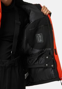 Giacca isolante arancione con interno nero, dotata di tasca con zip, etichetta di cura e cinturini regolabili. Design elegante con varie texture.