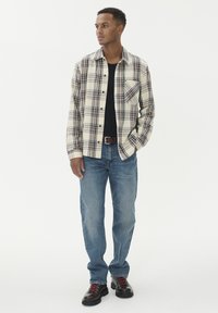 Karirana flanelasta srajca v krem, modri in burgundski barvi, oblečena čez črno majico, v kombinaciji z svetlo modrimi džins hlačami in temnimi črnimi čevlji na vezalke.