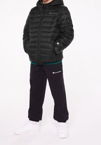 Czarna puchowa kurtka z zamkiem z przodu i kapturem, zestawiona z czarnymi dresami z logo oraz białymi sneakersami. Gładka faktura i casualowy styl.