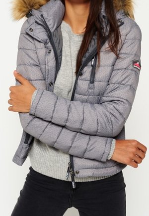 Frau trägt eine graue gesteppte Jacke mit pelzgefütterter Kapuze, teilweise über einen hellgrauen Pullover gezippt, Arme vor der Brust verschränkt.