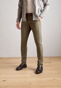Pantalon ajusté vert olive avec une texture lisse, associé à des chaussures slip-on noires ornées d'une boucle dorée. Porté avec une chemise à motifs.