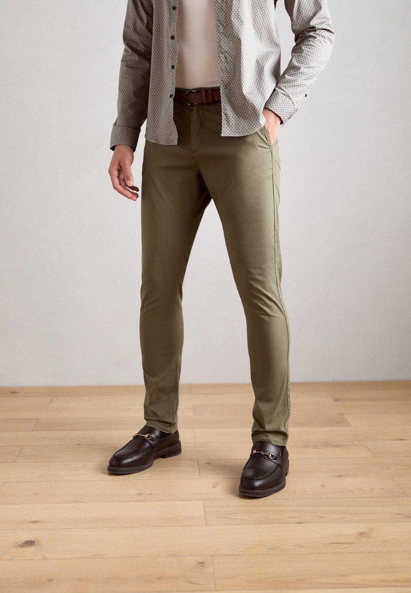 Pantalon ajusté vert olive avec une texture lisse, associé à des chaussures slip-on noires ornées d'une boucle dorée. Porté avec une chemise à motifs.