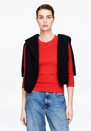 Femme portant un haut rouge à manches trois-quarts, un jean bleu, un pull noir posé sur les épaules et un fin collier argenté, debout devant un fond blanc.