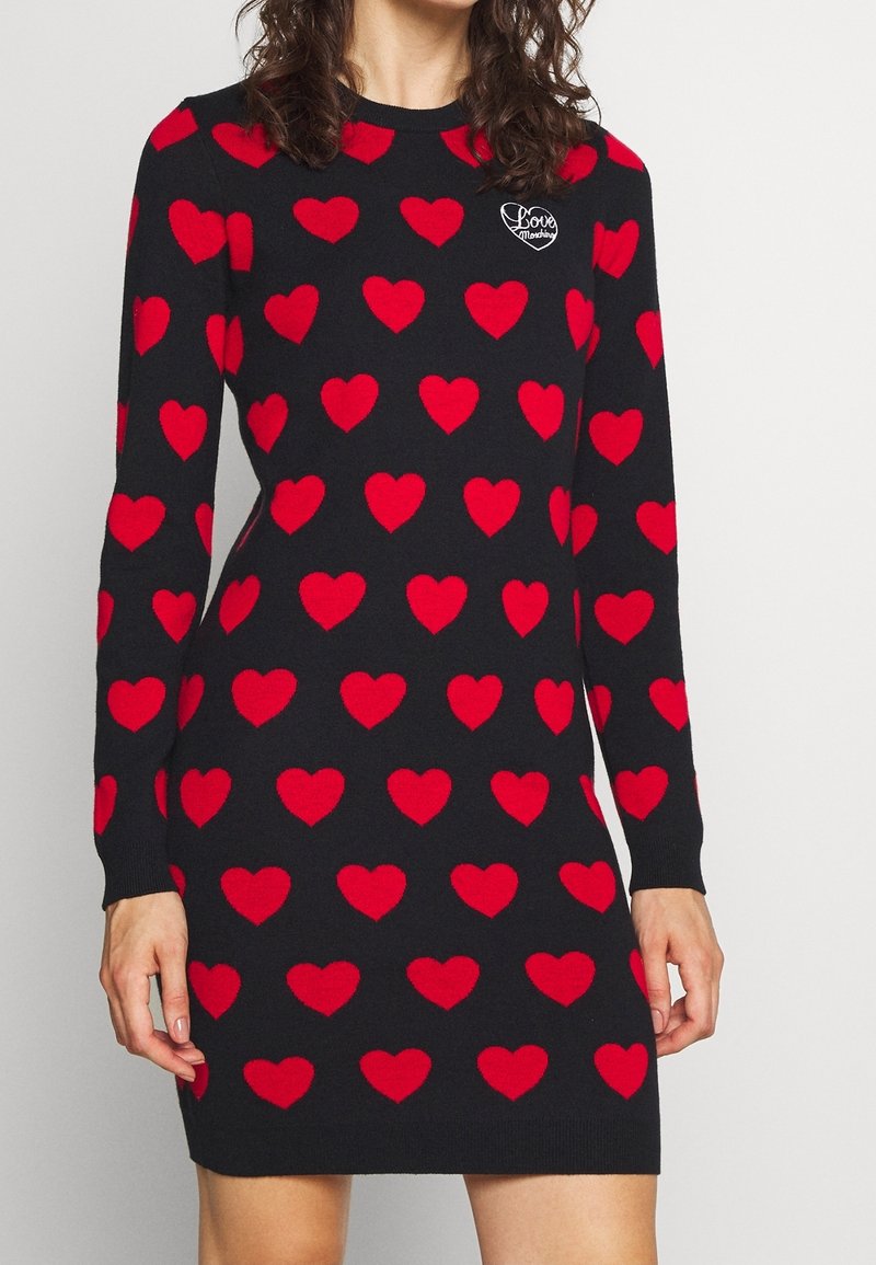 Robe noire à manches longues avec des motifs en forme de cœurs rouges et un petit logo blanc en forme de cœur « love » sur la poitrine portée par une femme.