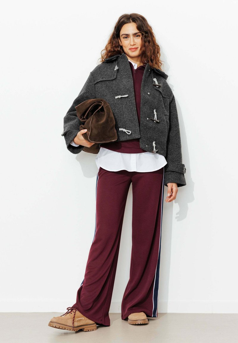 Veste en laine grise cintrée avec des fermetures à bascule, pantalon de sport bordeaux avec des rayures sur les côtés, et chaussures à lacets beige, tenant un sac en tissu marron.