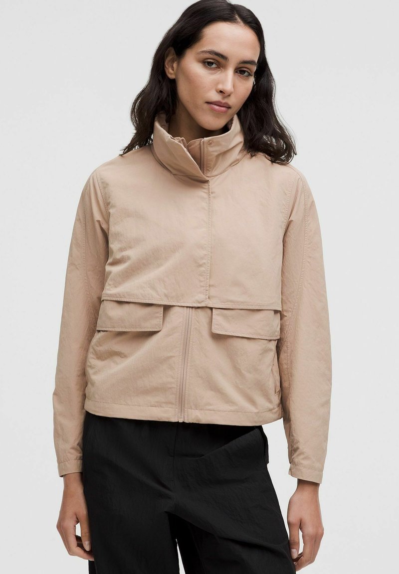 Femme portant une veste zippée beige avec un col montant et des poches à rabat, associée à un pantalon noir ample, se tenant devant un fond uni.