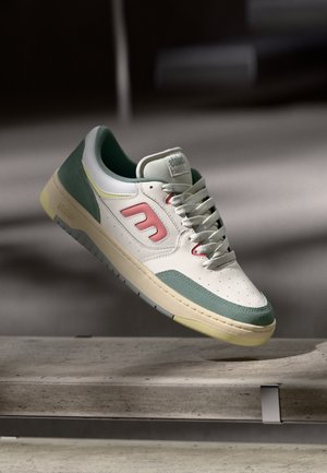 Witte sneaker met groene en roze accenten, witte veters en een beige zool, zwevend boven een betonnen oppervlak in zacht licht.
