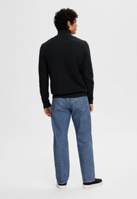 Black knit turtleneck sweater, fitted, worn with light blue straight-leg jeans and dark footwear, viewed from the back.