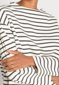 Persona que lleva una camisa blanca de manga larga con rayas negras horizontales, con los brazos cruzados, cortada a la altura del cuello y la cintura.