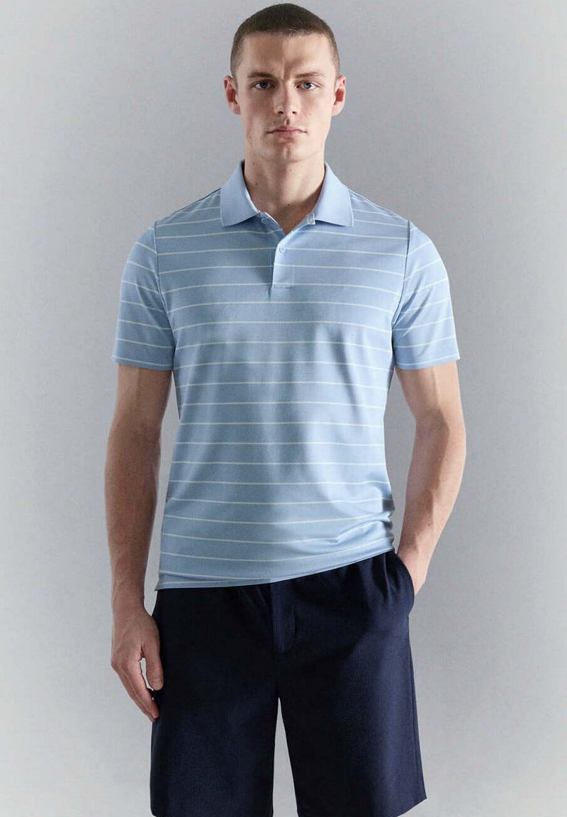 Jeune homme portant un polo rayé bleu clair et un short bleu marine foncé, debout devant un fond uni gris clair.