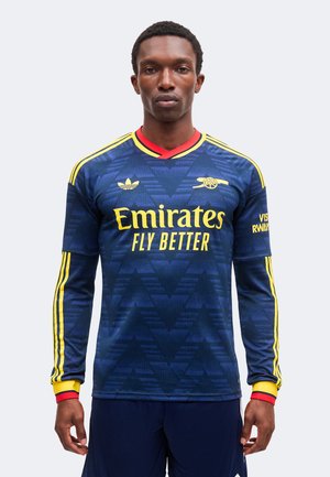 Homme portant un maillot de football Arsenal bleu foncé à manches longues avec des bandes jaunes, un logo jaune Emirates Fly Better et une bordure de col rouge, faisant face.