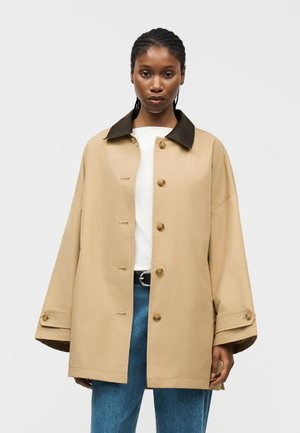 Donna che indossa un cappotto beige oversize con collo scuro e bottoni, top bianco, jeans blu, in piedi su uno sfondo chiaro uniforme.