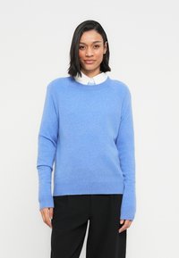 Pull bleu clair en matière douce, avec manches longues, encolure arrondie et coupe décontractée, associé à un pantalon noir.