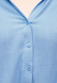 Ανοιχτό γαλάζιο πουκάμισο από υφασμάτινη ύφανση με τρία κουμπιά κλειστά, εμφανίζοντας ελαφρά ζάρωμα κοντά στο πάνω κουμπί και V-σχήμα λαιμόκοψη.