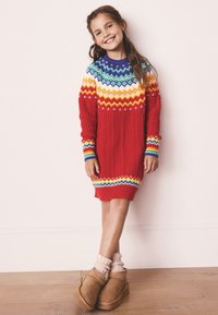 Červené pletené svetrové šaty s barevným vzorovaným klínem. Má strukturu copánkového pletení a žebrovaný lem. Kombinováno s béžovými kotníkovými botami a světlými ponožkami.