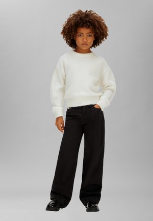 Pull blanc duveteux avec poignets et ourlet côtelés, associé à un pantalon noir à coupe ample et des chaussures noires brillantes, debout devant un fond gris.