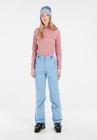 Lichtblauwe skibroek met hoge taille, ritszakken en een rode langärmige top, gecombineerd met een grijs en blauwe beanie. Zwarte skischoenen.