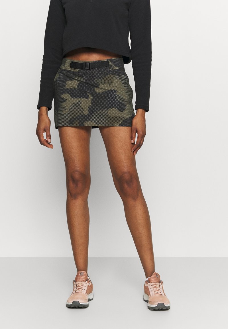 Minifalda de camuflaje con textura de malla, ceñida en la cintura con un cinturón. Combinada con zapatillas deportivas de color rosa claro, sobre un fondo liso.