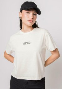 Camiseta blanca corta con mangas cortas y el texto "CRUSHING MACHINE" en negro. Presenta una textura de algodón suave, combinada con una gorra negra.