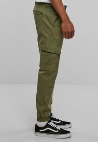 Olijfgroene cargobroek met meerdere zakken, elastische manchetten en een zachte textuur, gecombineerd met zwarte sneakers met witte accenten.