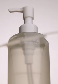 Przezroczysta plastikowa butelka oleju do oczyszczania z białą pompką. Etykieta głosi "Organiczne Kwiaty". Gładka tekstura, półprzezroczysta obudowa, zaokrąglony kształt.