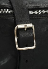 Detail van een zwarte leren tas met een zilveren metalen gesp op een gestructureerde schouderband, met een rits zichtbaar op de achtergrond.