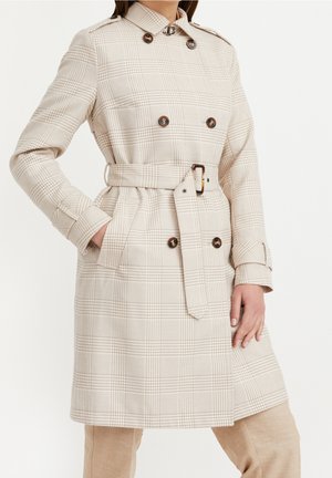 Femme portant un trench-coat ceinturé beige à carreaux avec des boutons croisés et des poches latérales, posant avec une main dans la poche.