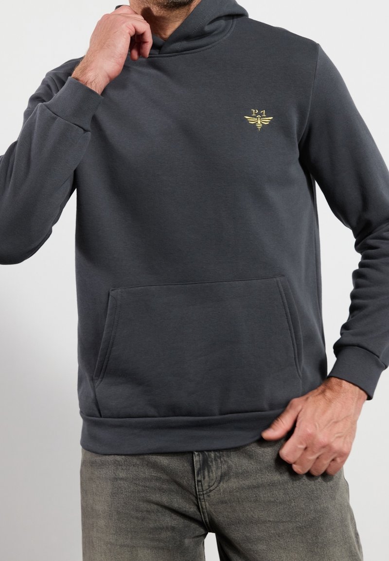 Hombre con sudadera gris oscuro con bolsillo frontal y pequeño logo de insecto amarillo en el pecho, combinada con jeans grises.