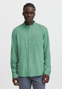 Grøn langærmet skjorte med bandkrave, frontlukning med knapper, enkelt brystlomme og let struktur. Bæres med grå jeans.