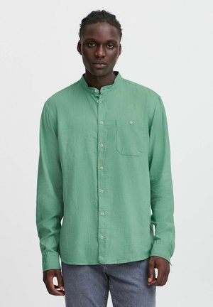 Camicia verde a maniche lunghe con colletto a fascia, chiusura frontale con bottoni, taschino singolo sul petto e leggero texture. Indossata con jeans grigi.