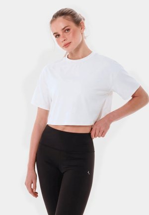 Młoda kobieta z włosami związanymi do tyłu, ubrana w biały krótki t-shirt i czarne legginsy z wysokim stanem, stoi z jedną ręką na biodrze.