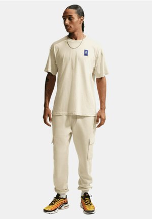 T-shirt in cotone beige con una toppa grafica blu, abbinata a pantaloni cargo beige con tasche e scarpe da ginnastica multicolori.