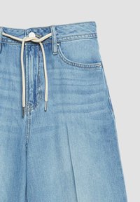 Hellblauer Jeansrock mit Vordertaschen, Gürtelschlaufen, weißem Knopf und einem beigen Kordelzug, der an der Taille gebunden ist.