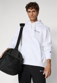White hoodie with a front pocket, featuring a black embroidered logo. Black bag with mesh panel and adjustable strap. Black pants with logo.